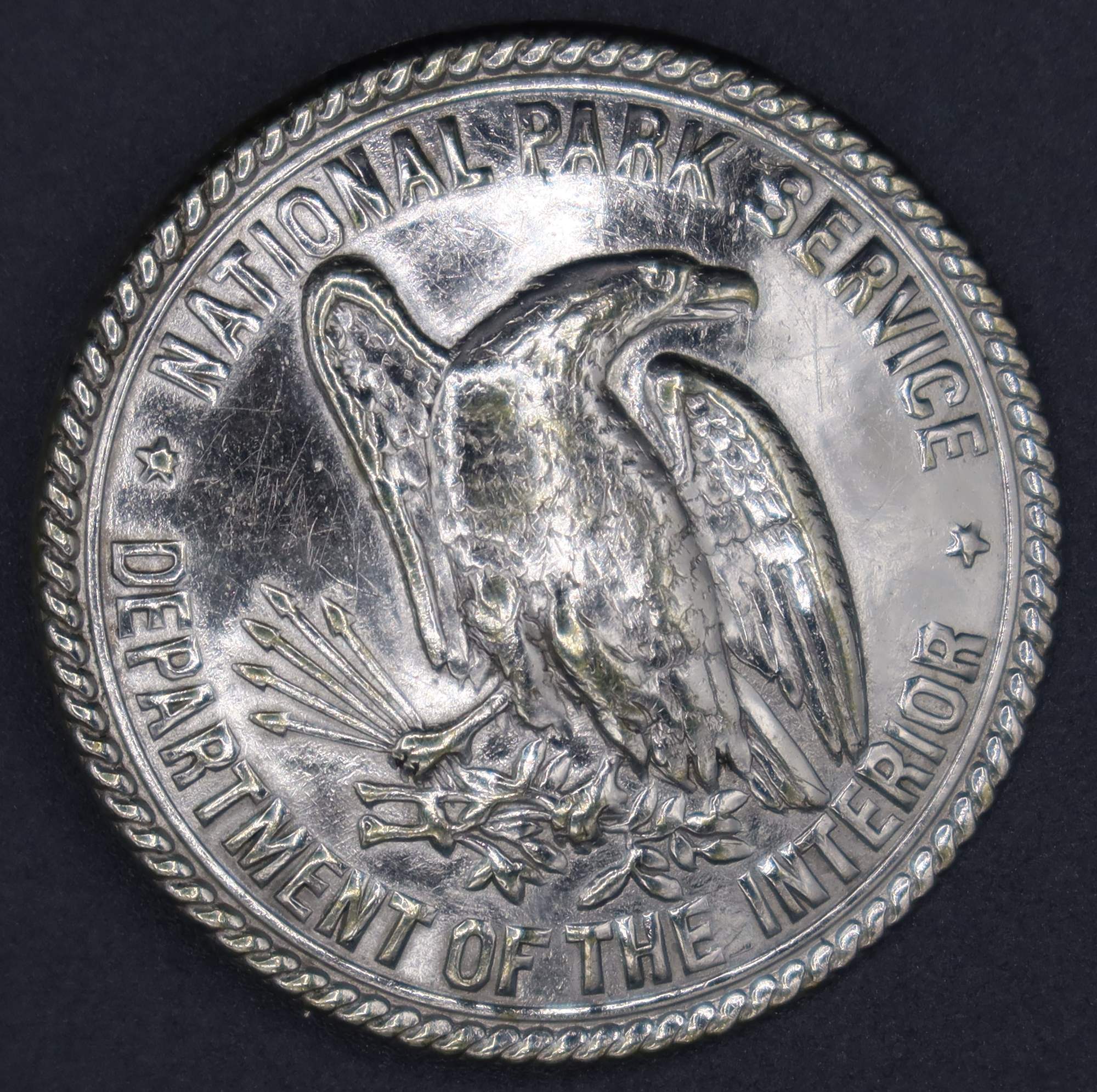 Round silver badge with an eagle in the middle center, marked National Park Service, Department of the Interior. 